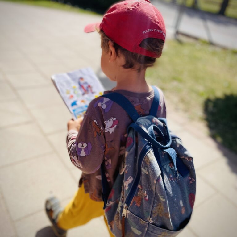 Preschooler with backpack and red baseball cap walking across a sunny yard.